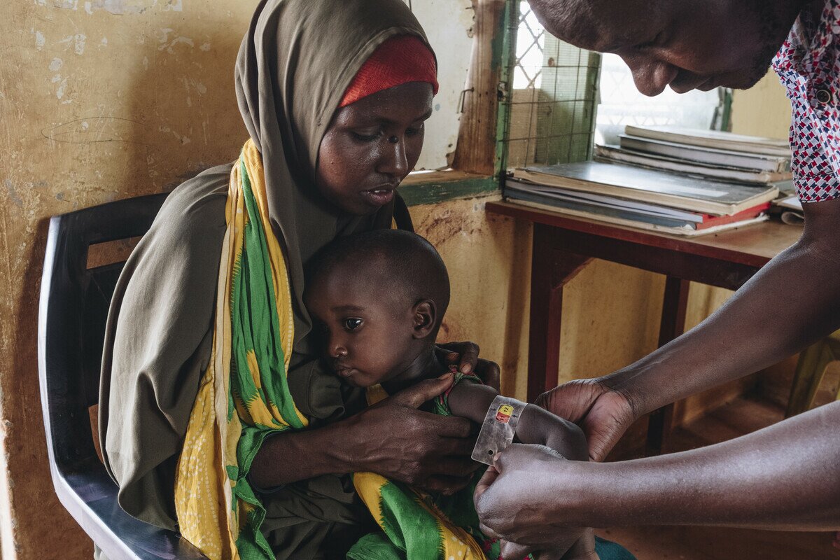 Mother holding her young child as a man measures the child's arm with a mid-upper arm circumference measuring tape.